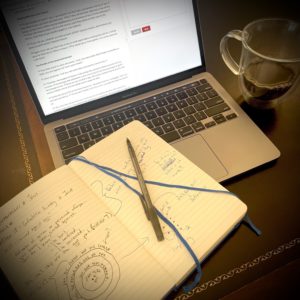 A desk, featuring a notebook and pen resting on an open laptop next to a cup of coffee.
