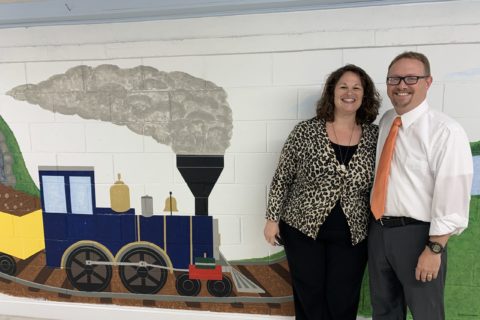 A woman wearing a nice cardigan standing and black shirt and pants standing next to a man with a suit and orange tie. Behind them is a mural with a train.