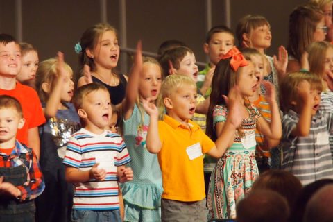 Children stand on stage in a line and sing and raise their hands