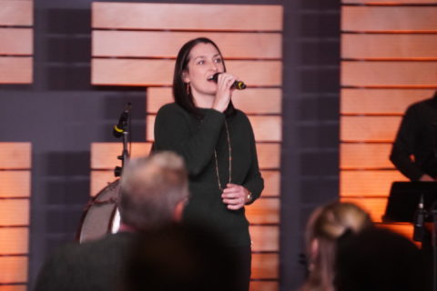 A young woman wearing all black sings on stage with a cross in the background. Boones Creek Christian Church