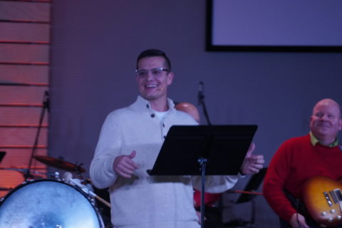 A man preaches behind a music stand with a band behind him. Boones Creek Christian Church