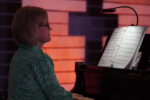 A woman sits at the piano in the dark with just the music lit and the cross in the background at Boones Creek Christian Church