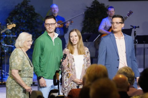 A preacher with a suit coat stands beside a young couple dressed in Sunday clothes and an older woman beside them. All four are on stage. Boones Creek Christian Church