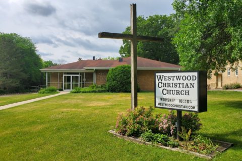A bright green lawn with a cross in front and a sign that reads Westwood Christian Church. A small brown church building in the background.