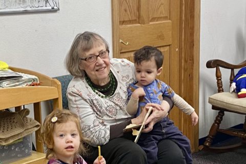 An older women plays with two children in a nursery. A bright yellow kids table is in front of them.