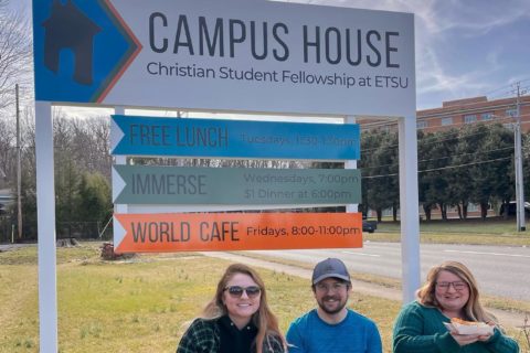 Three students sit in front of a sign that reads Campus House